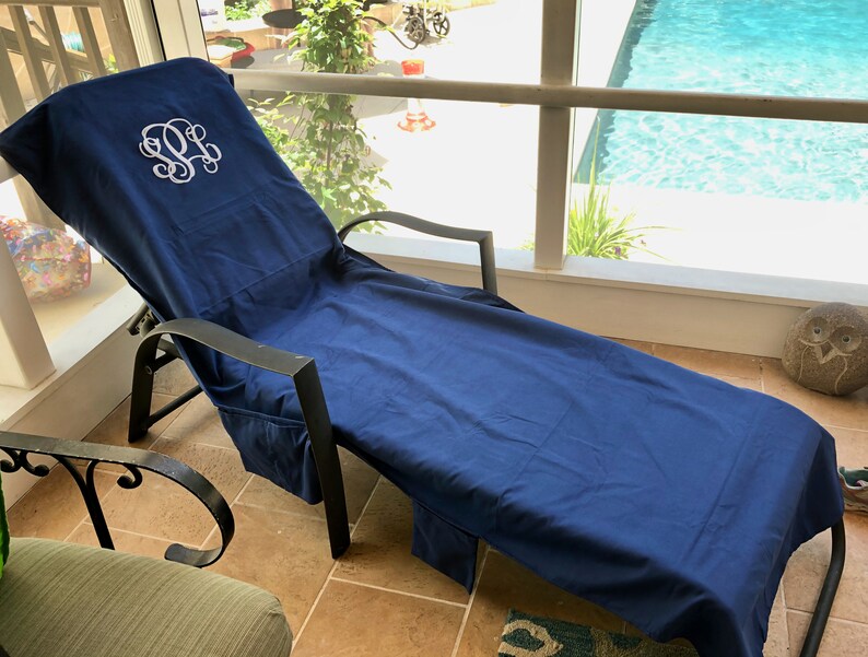 A navy blue microfiber sueded pool chair cover is draped completely over a lounge chair by a pool. It has a white monogram where someone’s head will lay, indicating these can be monogrammed or personalized with a name.