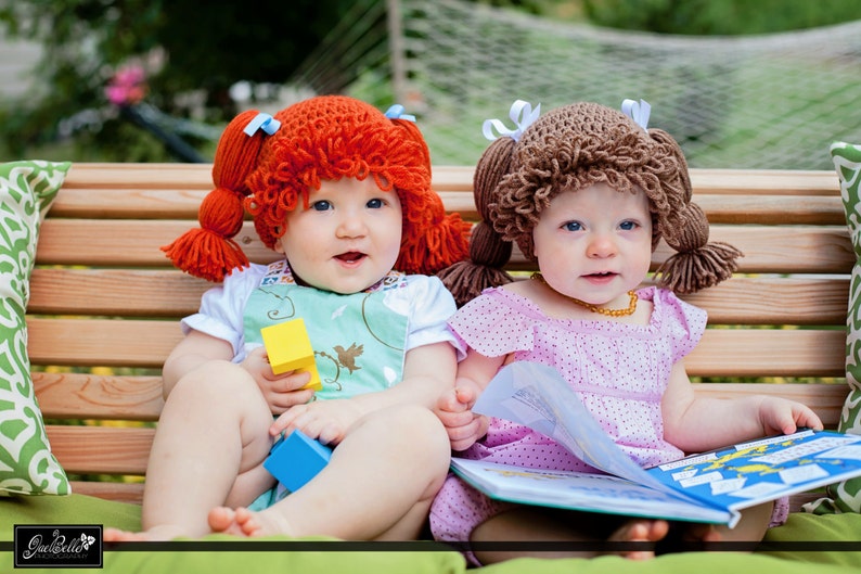 Two baby girls are sitting on a bench playing with toys and wearing the Cabbage Patch crochet hats. Ones wearing a pumpkin orange (now retired) hat and the other is  light brunette hat color.