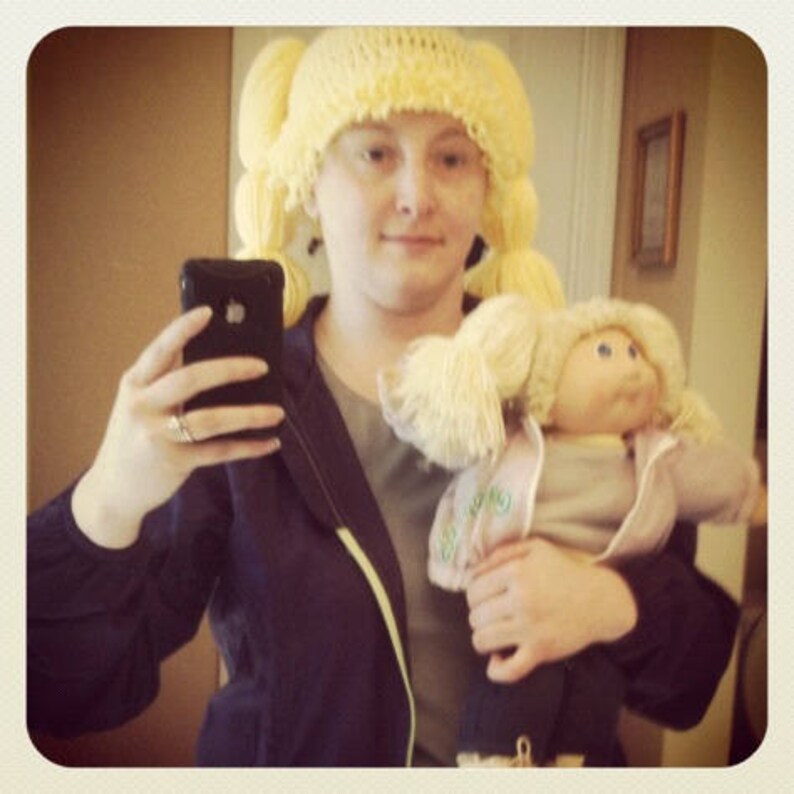 A woman is wearing a pale blonde adult sized crochet Cabbage Patch hat with purple bows. It matches the hair of the vintage 1980s Cabbage Patch Doll she is holding in her arms.