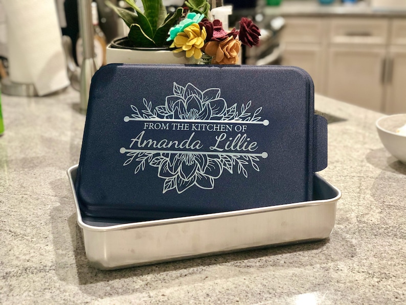 An aluminum cake pan with a navy blue lid sits on a grey marble kitchen counter, showing that the navy lid is laser engraved with a Magnolia design and the words From the Kitchen of and a name, indicating that these are personalized.