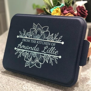 An aluminum cake pan with a navy blue lid sits on a grey marble kitchen counter, showing that the navy lid is laser engraved with a Magnolia design and the words From the Kitchen of and a name, indicating that these are personalized.