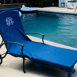 A navy blue microfiber sueded pool chair cover is draped completely over a lounge chair by a pool. It has a white monogram where someone’s head will lay, indicating these can be monogrammed or personalized with a name.