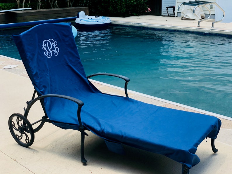 A navy blue microfiber sueded pool chair cover is draped completely over a lounge chair by a pool. It has a white monogram where someone’s head will lay, indicating these can be monogrammed or personalized with a name.