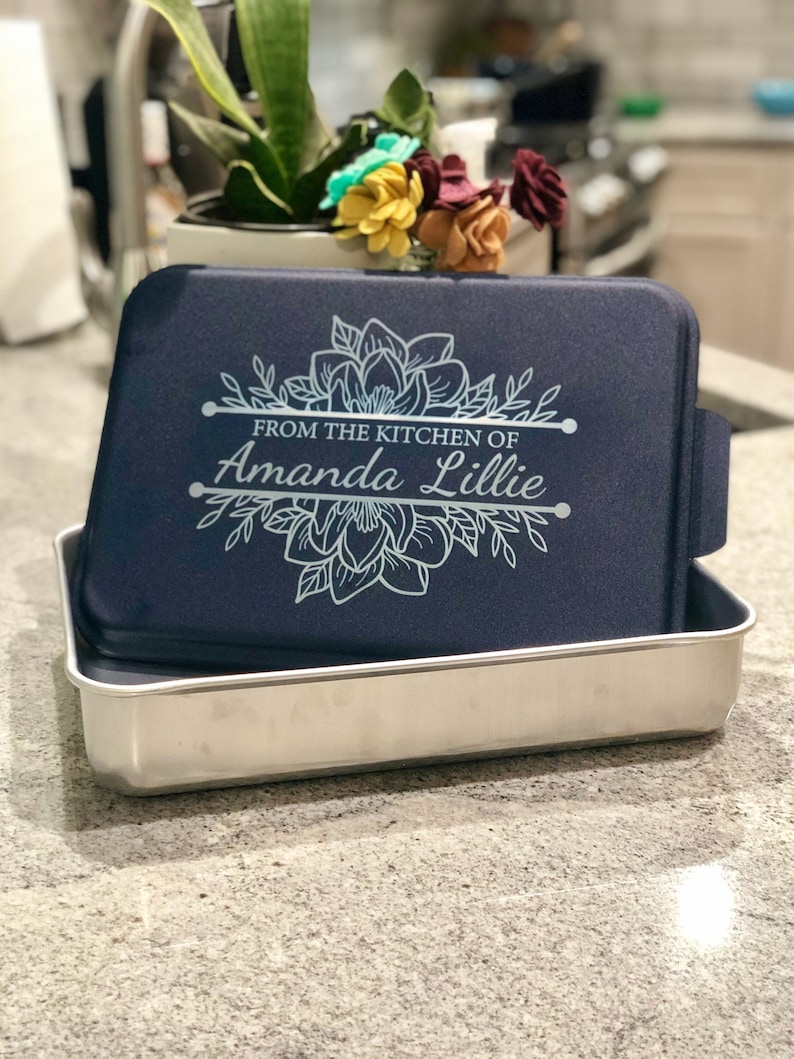 An aluminum cake pan with a navy blue lid sits on a grey marble kitchen counter, showing that the navy lid is laser engraved with a Magnolia design and the words From the Kitchen of and a name, indicating that these are personalized.