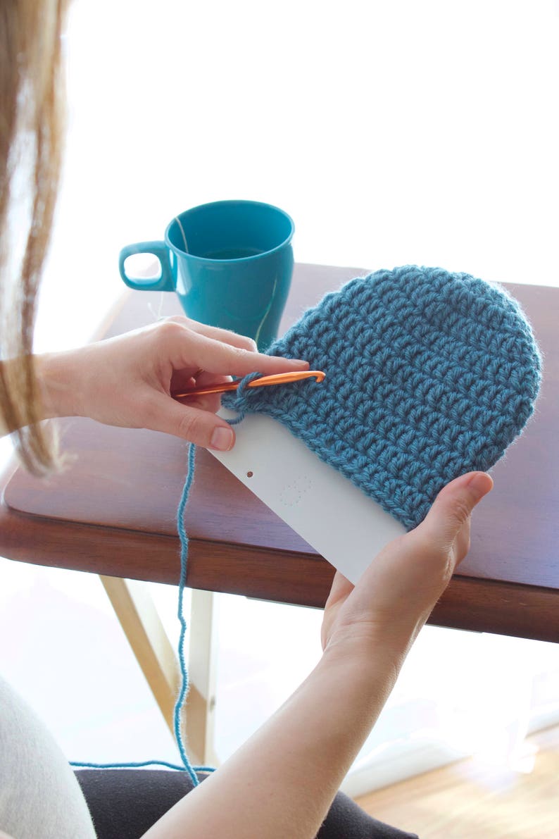 A lady is holding a partially crocheted blue beanie hat that is slipped over the template to show how far she needs to crochet before the hat is the right length she needs it to be.