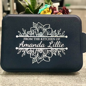 An aluminum cake pan with a navy blue lid sits on a grey marble kitchen counter, showing that the navy lid is laser engraved with A Magnolia design and the words From the Kitchen of and a name, indicating that these are personalized.