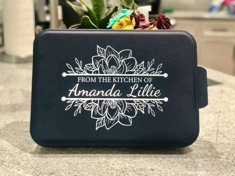 An aluminum cake pan with a navy blue lid sits on a grey marble kitchen counter, showing that the navy lid is laser engraved with A Magnolia design and the words From the Kitchen of and a name, indicating that these are personalized.