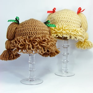 Two finish cabbage patch crochet hats sit on hand stands, showing the looped bangs and 2 pigtails with bows on top of the pigtails.