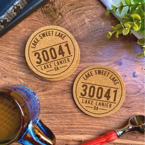 May include: Two round cork coasters with the text "Lake Sweet Lake 30041 Lake Lanier GA" printed on them. The coasters are on a wooden surface.