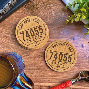 Op de afbeelding: Twee kurken onderzetters met de tekst "Home Sweet Home" en het adres "74055 Owasso OK" erop gedrukt. De onderzetters liggen op een houten oppervlak.