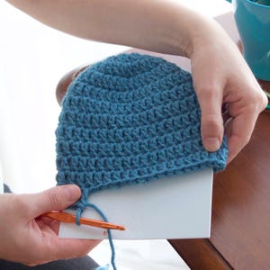 A lady is holding a partially crocheted blue beanie hat that is slipped over the template to show how far she needs to crochet before the hat is the right length she needs it to be.