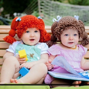 Two baby girls are sitting on a bench playing with toys and wearing the Cabbage Patch crochet hats. Ones wearing a pumpkin orange (now retired) hat and the other is  light brunette hat color.