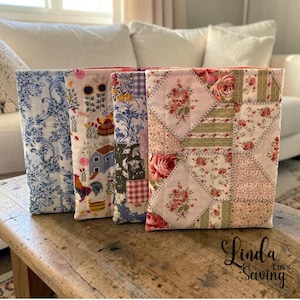 May include: Four rectangular fabric book covers featuring various patterns including blue and white toile, a farm scene with roosters and sunflowers, a patchwork design with floral prints, and a pink floral quilt pattern with stitched details.