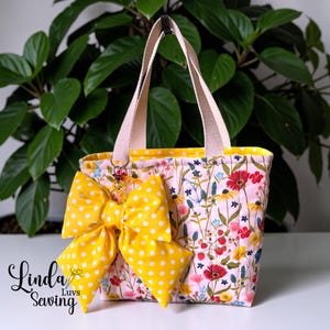 May include: A floral print tote bag with a large yellow polka dot bow. The bag features a pink background with colorful flowers and a yellow interior. The handles are beige.