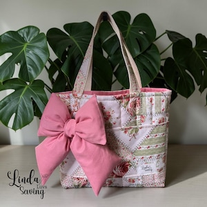 May include: A quilted tote bag with a large pink bow. The bag features a patchwork design with floral and striped patterns in pink, green, and white. The handles are beige. The text "Linda Luvs Saving" is visible.