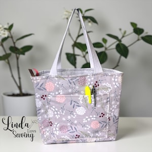 May include: A floral tote bag with a light gray background, featuring a pattern of pink and white flowers and green leaves. The bag has a front pocket with a yellow pen and a black pen. The handles are white.