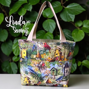 May include: A fabric tote bag with a colorful birdhouse and bird print. The bag has beige handles and a matching interior. The text "Linda Luvs Saving" is printed on the upper left corner of the bag.