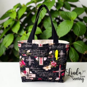 May include: Black tote bag with a floral and cross pattern, featuring the text "Amazing Grace." The bag has black handles and a pocket with a pen. The interior lining is a cream color.