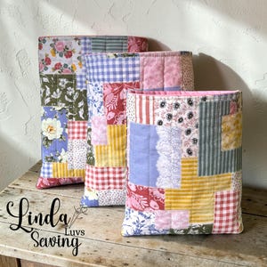 May include: Three patchwork fabric containers in a row, each with a unique mix of floral, striped, and gingham patterns in various colors. The containers are on a wooden surface, with the text "Linda Luvs Sewing" in the lower left corner.