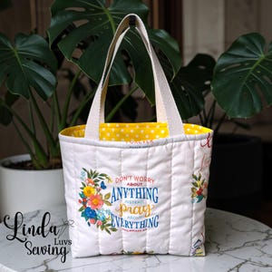 May include: White quilted tote bag with floral accents and the text "Don't worry about anything, instead, pray about everything." The interior is yellow with white polka dots. The bag has cream-colored handles.