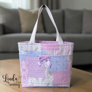 May include: A quilted tote bag featuring a patchwork design in shades of lavender, light blue, and white with floral, gingham, and polka dot patterns. The bag has two white fabric handles and a small, centered lavender satin ribbon bow.