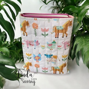 May include: A white quilted fabric tote bag with a farm animal pattern, including horses, pigs, and cows, and floral accents. The bag has a pink interior and is surrounded by green foliage. The text "Linda Luvs Sewing" is visible on the lower left.