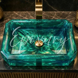 May include: A rectangular vessel sink made of translucent emerald green resin with a swirling pattern, featuring a central gold-toned drain cover. The sink is paired with a matching gold-toned faucet and is set against a dark marble countertop.