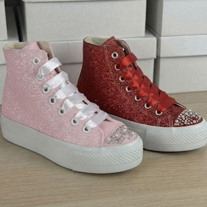 May include: Pink and red glitter high-top sneakers with white soles. The pink sneakers have white ribbon laces and the red sneakers have red ribbon laces. Both pairs have decorative rhinestones on the toe.
