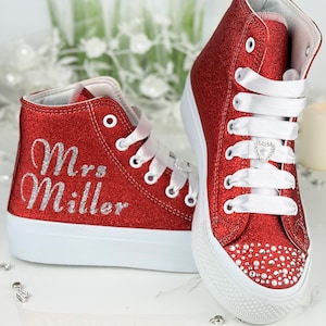 Puede incluir: Zapatillas altas rojas con purpurina, suelas y cordones blancos. Las zapatillas tienen el nombre "Mrs. Miller" en purpurina plateada en el lateral. La puntera del zapato está adornada con pequeños cristales y los cordones con un charm en forma de corazón.