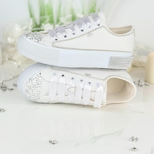 Könnte beinhalten: Zwei weiße Leinenschuhe mit Strass- und Bandakzenten. Die Schuhe haben weiße Gummisohlen und sind mit Strasssteinen auf Zehen und Ferse verziert. Die Schnürsenkel sind weiße Satinbänder.