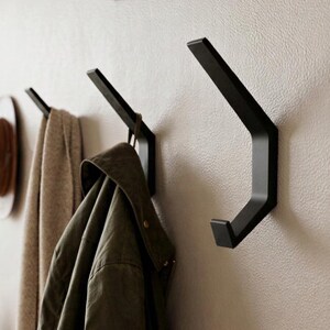 May include: Three matte black wall-mounted hooks with an angular, geometric design. The hooks are attached to a textured wall, with a brown hat, a beige scarf, and an olive green jacket hanging from the two hooks on the left side of the frame.