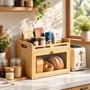 May include: A bamboo bread box with a fluted glass door, containing a loaf of sliced bread, a round loaf, and a bagel. The top shelf holds two paper bags, two glass jars, a blue metal can, and a white bag of cookies.