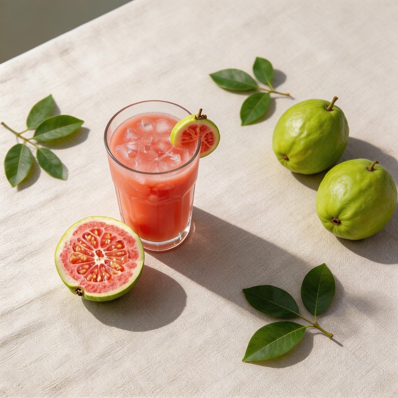 May include: A clear glass filled with pink guava juice and ice cubes, garnished with a slice of pink guava. Beside the glass sits a halved pink guava, two whole green guavas, and several green leaves arranged around the beverage.