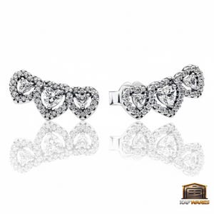 May include: A pair of silver-toned stud earrings. Each earring features three heart-shaped designs, each surrounded by small, clear crystals. The earrings are displayed on a white surface, with their reflections visible.