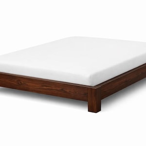 May include: A low-profile wooden bed frame with a dark brown finish. The bed features a simple, rectangular design with a white mattress. The frame is made of solid wood and has a minimalist aesthetic.