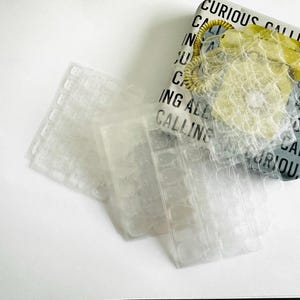 Peut inclure: Des autocollants d'art d'ongle adhésifs transparents et rectangulaires à côté d'une boîte décorative. La boîte a un fond gris et blanc avec les mots "CURIOUS CALLING ALL" imprimés. Un téléphone jaune est sur le dessus.