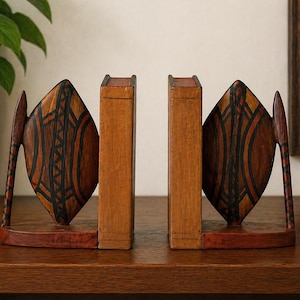 Peut inclure: Paire de serre-livres en bois avec un motif en forme de bouclier. Les serre-livres sont bruns avec des motifs géométriques noirs et orange. Ils sont posés sur une surface en bois.