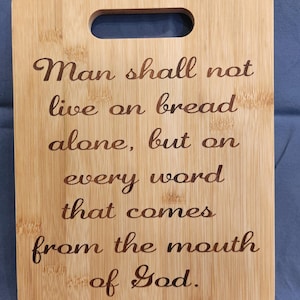 Op de afbeelding: Een rechthoekige bamboe snijplank met een handvat. De plank heeft gegraveerde tekst die luidt: "De mens zal niet van brood alleen leven, maar van elk woord dat uit de mond van God komt."