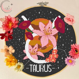 May include: A circular cross-stitch embroidery in a wooden hoop featuring a black background with white stars, a large orange planet, pink lily flowers, and the word TAURUS in white capital letters, decorated with various colorful artificial flowers.