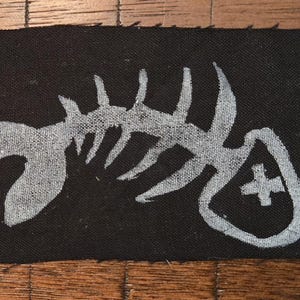 May include: Black rectangular patch featuring a white fish skeleton design. The patch has a raw edge and a cross symbol on the right. Likely for clothing or accessories, this patch is a unique design.