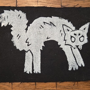 Peut inclure: Un écusson rectangulaire noir avec une illustration blanche d'un chat en position arquée. Le chat a de grands yeux, une queue touffue et une bouche légèrement ouverte, lui donnant un air effrayé ou défensif. L'écusson est probablement destiné aux vêtements ou aux accessoires.