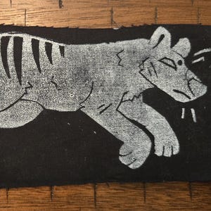 Op de afbeelding: Een rechthoekige, zwarte stoffen patch met een witte illustratie van een gestileerde buidelwolf, of Tasmaanse tijger, in een rennende pose. Het dier heeft strepen en een puntige snuit. De patch is waarschijnlijk handgemaakt.