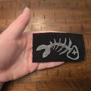 Pu&ograve; includere: Una toppa rettangolare nera con un disegno di scheletro di pesce bianco. La toppa, tenuta in mano, ha bordi grezzi. Lo scheletro di pesce &egrave; un'illustrazione dettagliata e stilizzata.