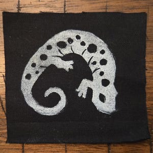 May include: A black square patch featuring a white, stylized gecko design. The gecko is depicted with a curved body, small limbs, and dotted patterns along its back. The design is hand-painted on the fabric.