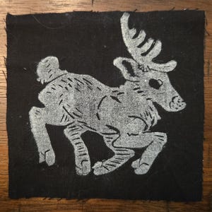 May include: A square, black fabric patch featuring a stylized, white deer design. The deer is depicted in a running pose with detailed antlers and body markings. The edges of the patch are unfinished.