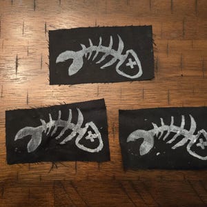 Pu&ograve; includere: Tre toppe di tessuto nero con un disegno di scheletro di pesce bianco. Ogni toppa presenta un'illustrazione dettagliata di uno scheletro di pesce con un simbolo a croce. Le toppe sono rettangolari e sembrano fatte a mano.