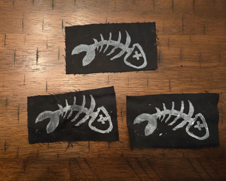 Pu&ograve; includere: Tre toppe di tessuto nero con un disegno di scheletro di pesce bianco. Ogni toppa presenta un'illustrazione dettagliata di uno scheletro di pesce con un simbolo a croce. Le toppe sono rettangolari e sembrano fatte a mano.