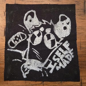 Pode incluir: Remendo quadrado preto com um design gráfico branco. O design apresenta formas abstratas e as palavras "SELF MADE" em uma fonte estilizada. O remendo é provavelmente para roupas ou acessórios.