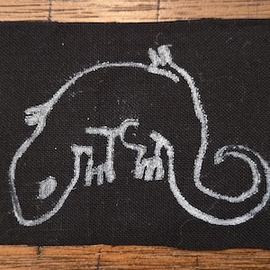 May include: Black fabric patch with a white, stylized drawing of a creature with a long tail and a rounded head. The creature's body has a series of rectangular shapes. The patch is rectangular and has a slightly frayed edge.