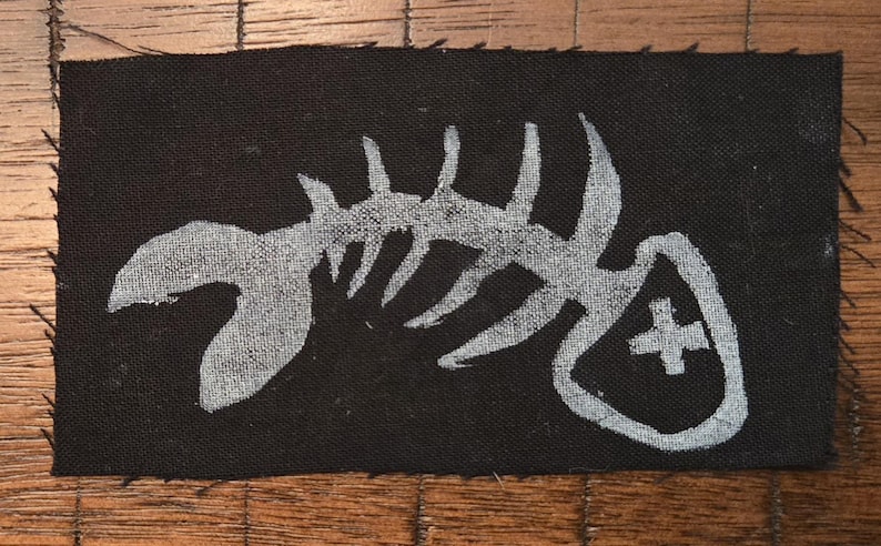 Pu&ograve; includere: Toppa rettangolare nera con un disegno di scheletro di pesce bianco. La toppa ha un bordo grezzo e un simbolo a croce sul lato destro. Adatta per abbigliamento o accessori.